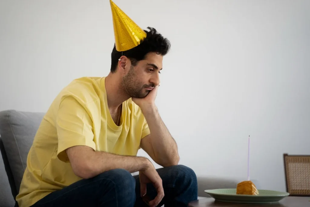 Homem com chapéu de festa, sentado sozinho, olhando para um bolo de aniversário, representando alguém que não gosta de comemorar seu aniversário.